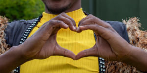 Person in a yellow shirt forms a heart shape with their hands, with a blurred outdoor background.